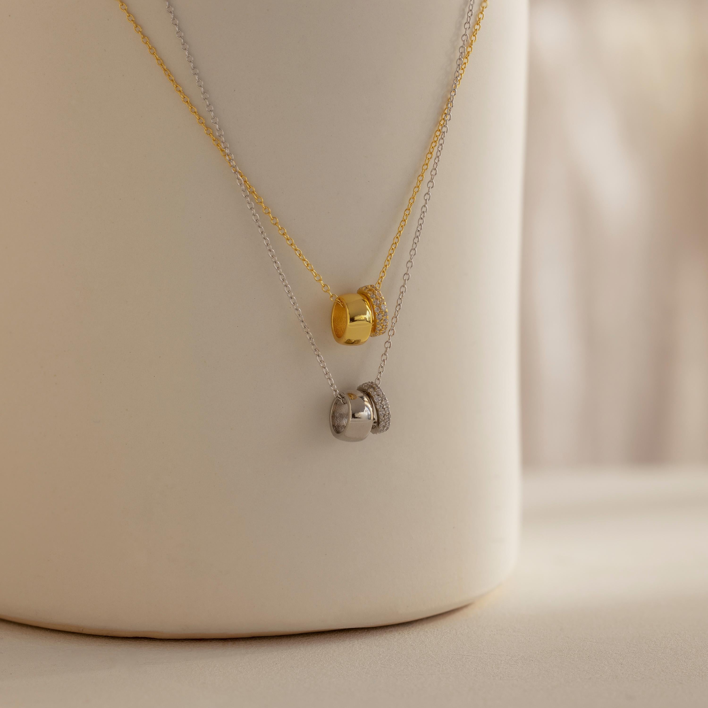 The Pave Duo Circle Necklace, featuring gold and silver pendants with pavé circles, is elegantly displayed on a white cylindrical stand—highlighting its refined beauty.