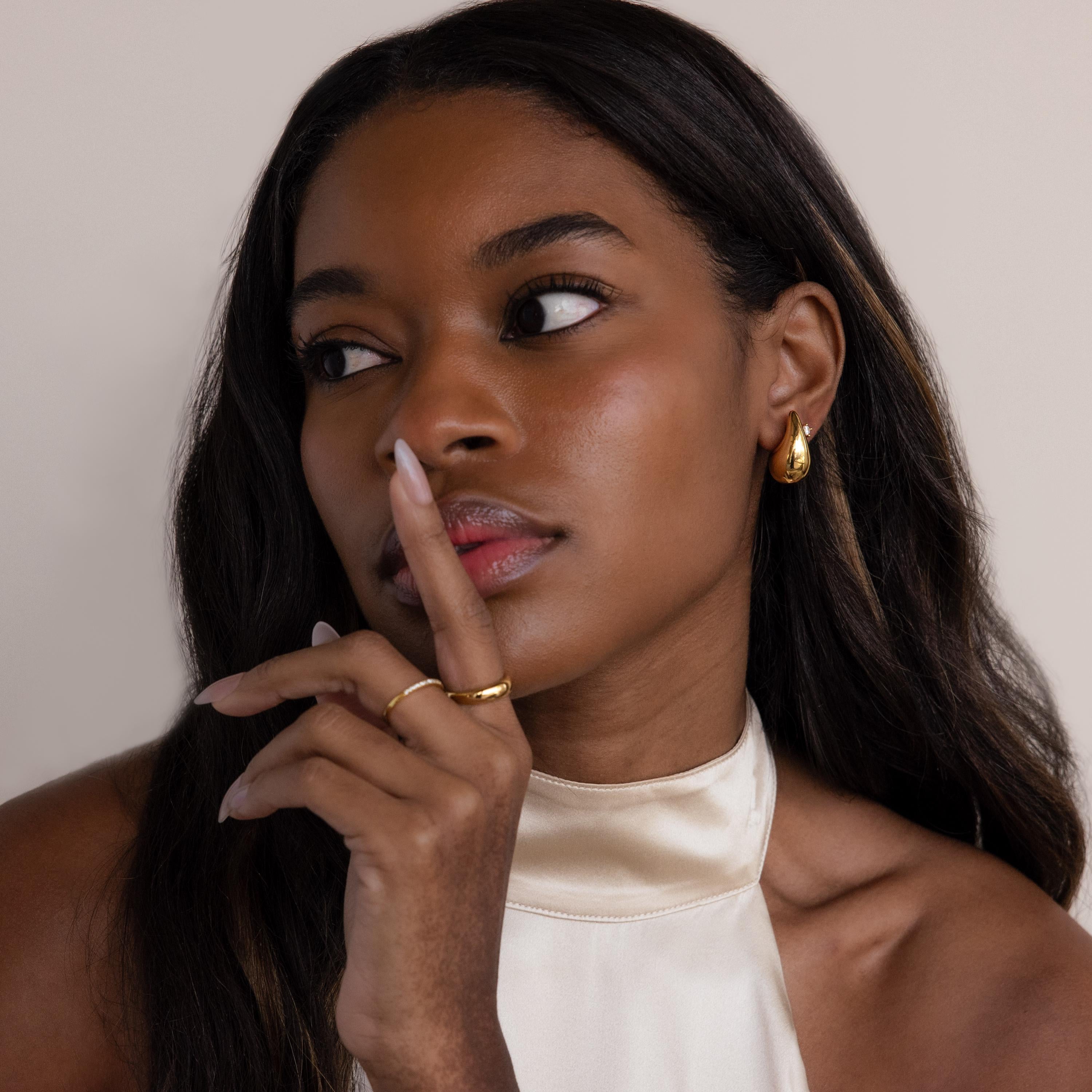 A woman with long dark hair wears the Chunky Tear Drop Studs gold earrings and a cream halter top, holding her finger to her lips.