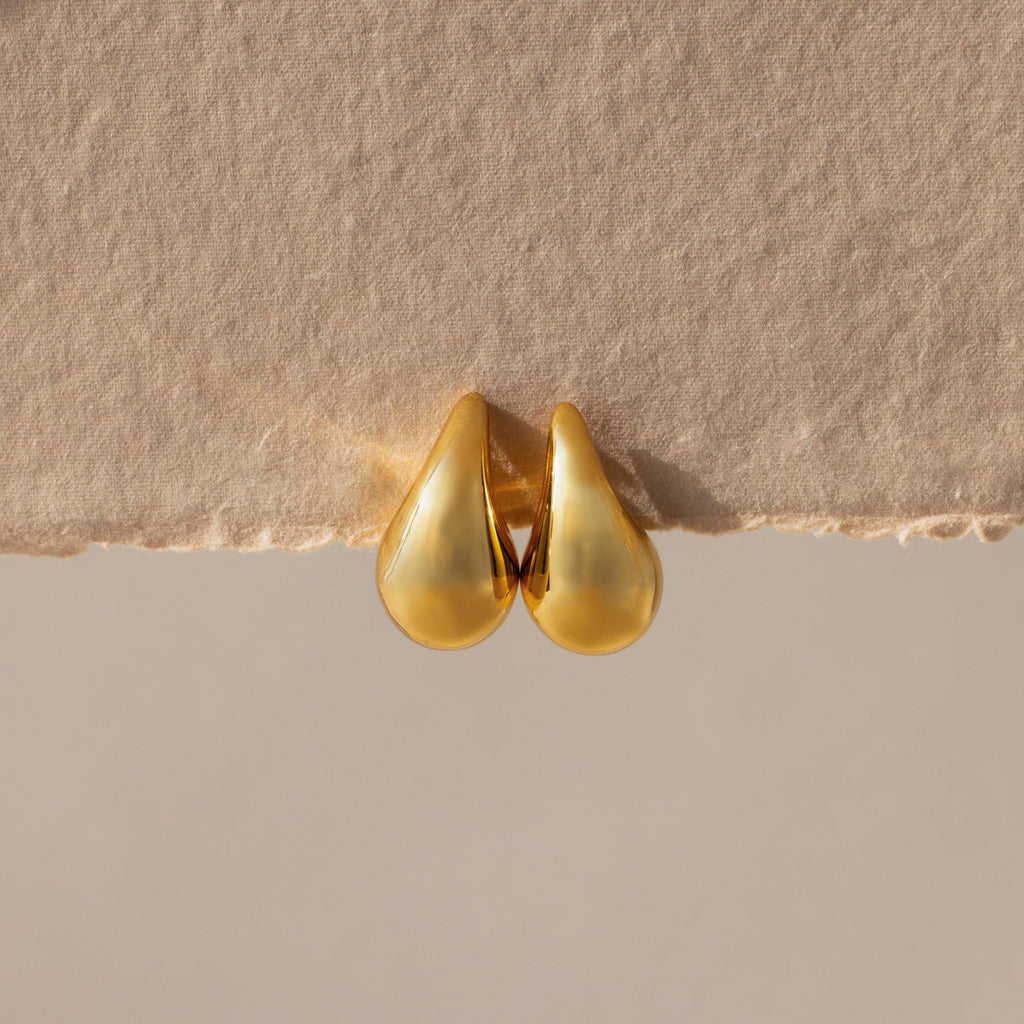 Chunky Tear Drop Studs rest on a textured beige surface, highlighting their bold silhouette. These oversized gold earrings make a statement and are perfect for elevating any outfit.