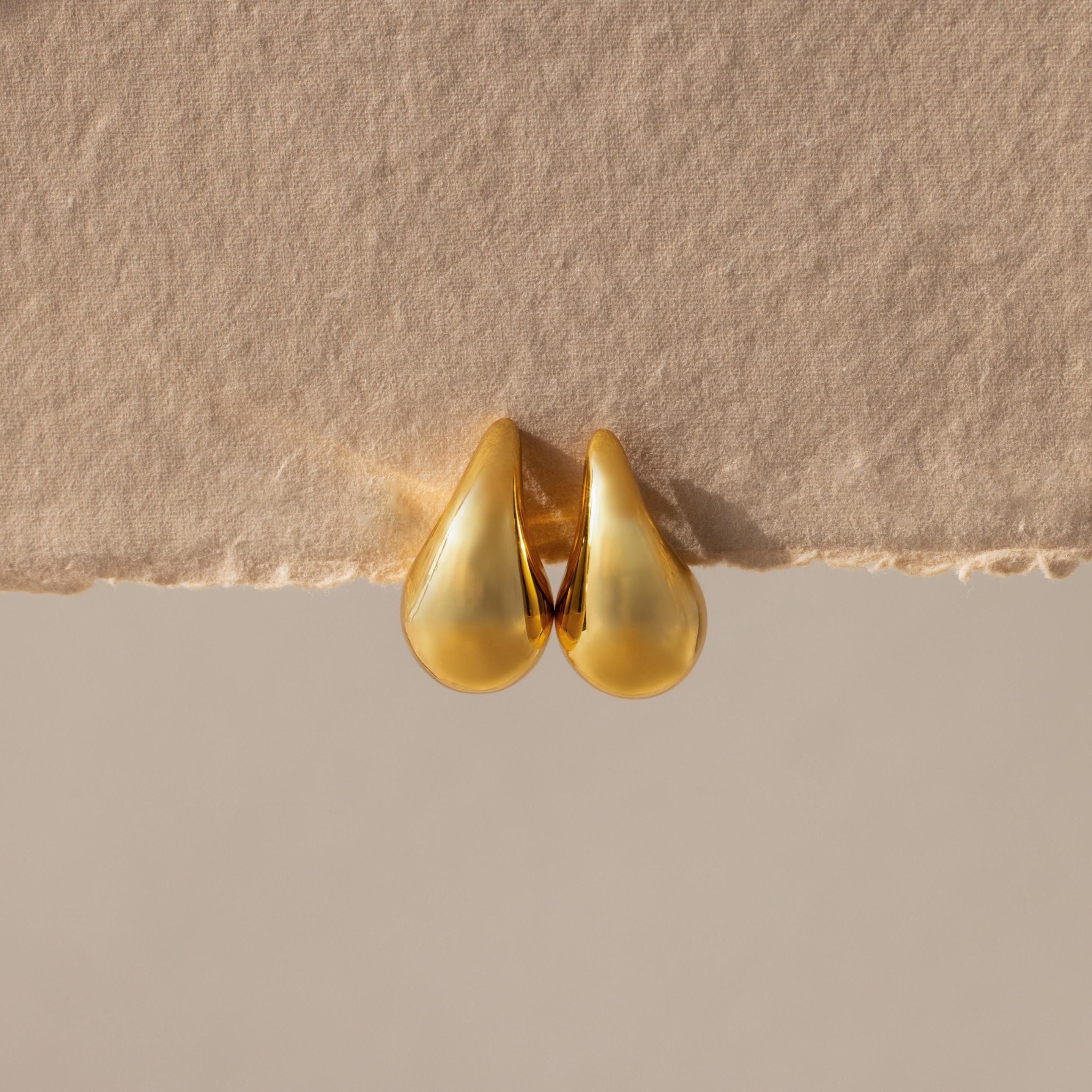 Chunky Tear Drop Studs rest on a textured beige surface, highlighting their bold silhouette. These oversized gold earrings make a statement and are perfect for elevating any outfit.