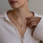 A woman in a cream sweater gently touches the elegant toggle clasp of the Paperclip Pearl Toggle Necklace, featuring a gold chain and a pearl pendant.