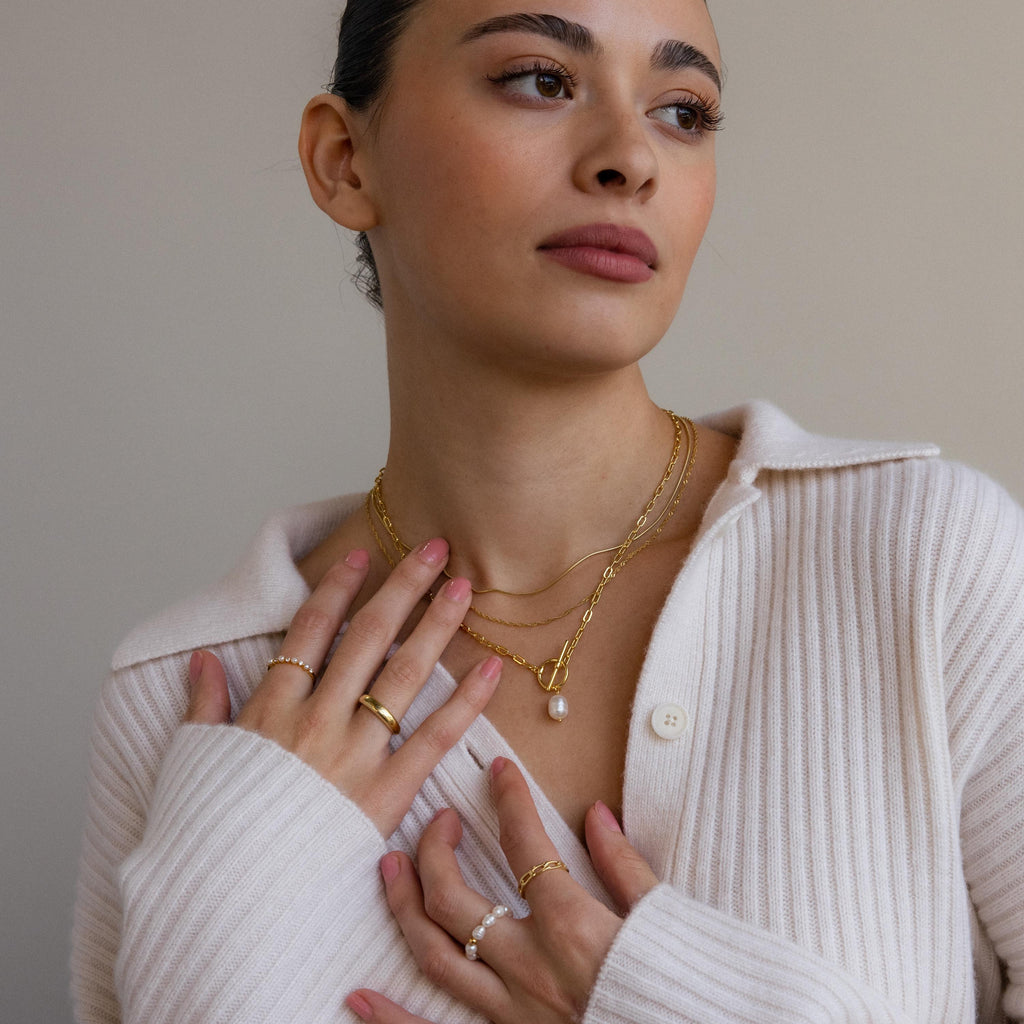 A woman in a cream ribbed sweater, looking away, wears layered gold necklaces and rings, featuring the Paperclip Pearl Toggle Necklace with a pearl pendant.
