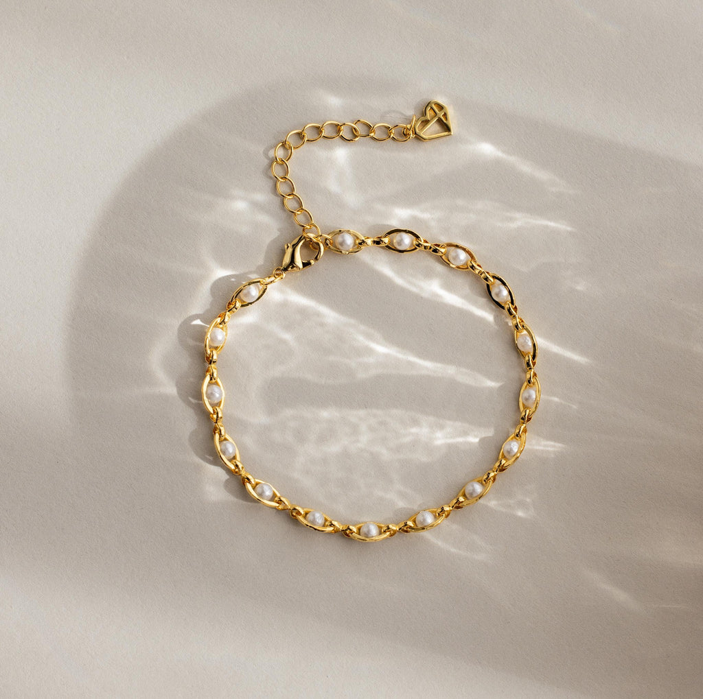 Eternity Pearl Bracelet: This elegant piece showcases pearls on a gold infinity chain, beautifully displayed flat on a beige surface with gentle, soft light reflections.