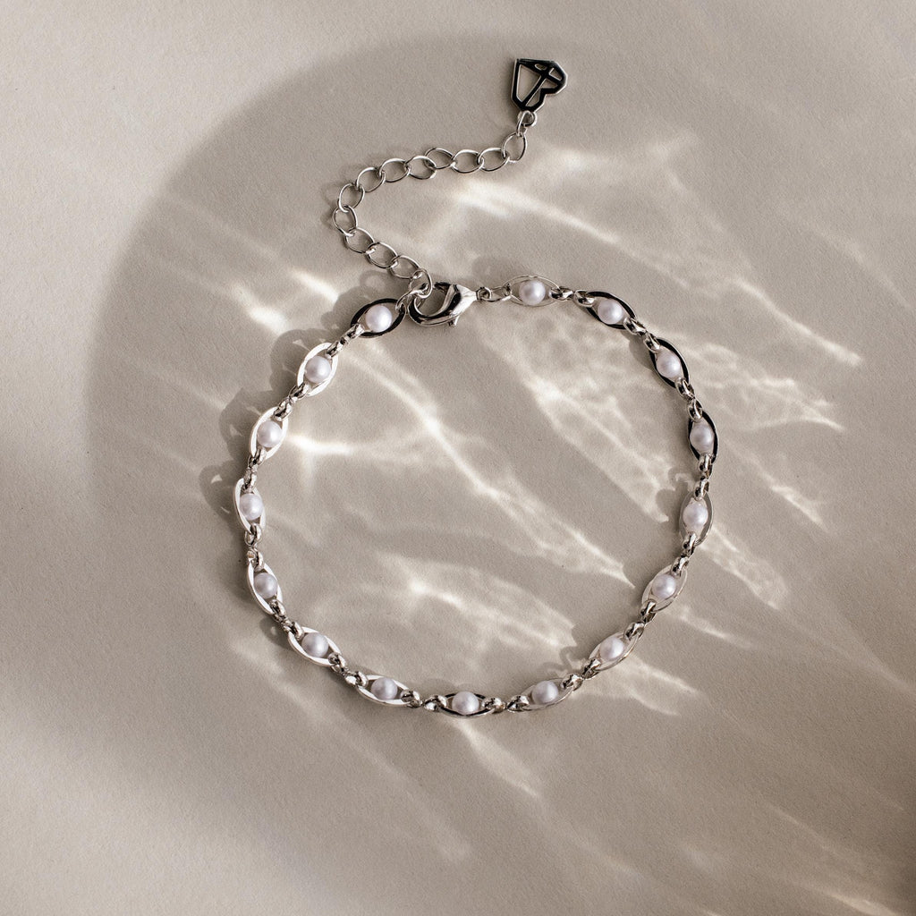 The Eternity Pearl Bracelet in Sterling Silver features a silver infinity chain with white pearls. Displayed on a neutral surface, its graceful charm is enhanced by soft light reflections, merging elegance with timeless style.