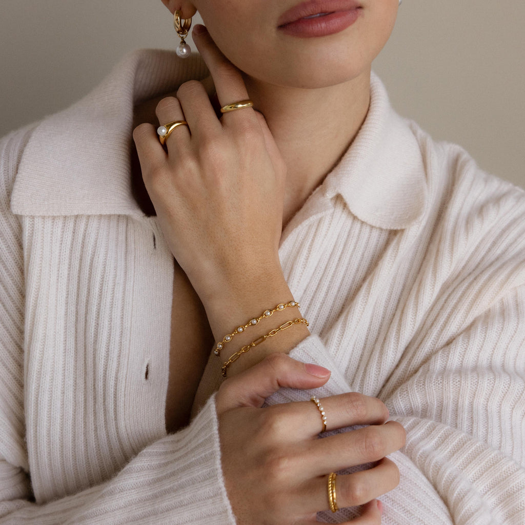 A woman in a cream sweater displays elegant jewelry—gold rings, bracelets, and a pearl earring—her hands near her face. The Eternity Pearl Bracelet and an infinity chain add the finishing touch to her sophisticated look.
