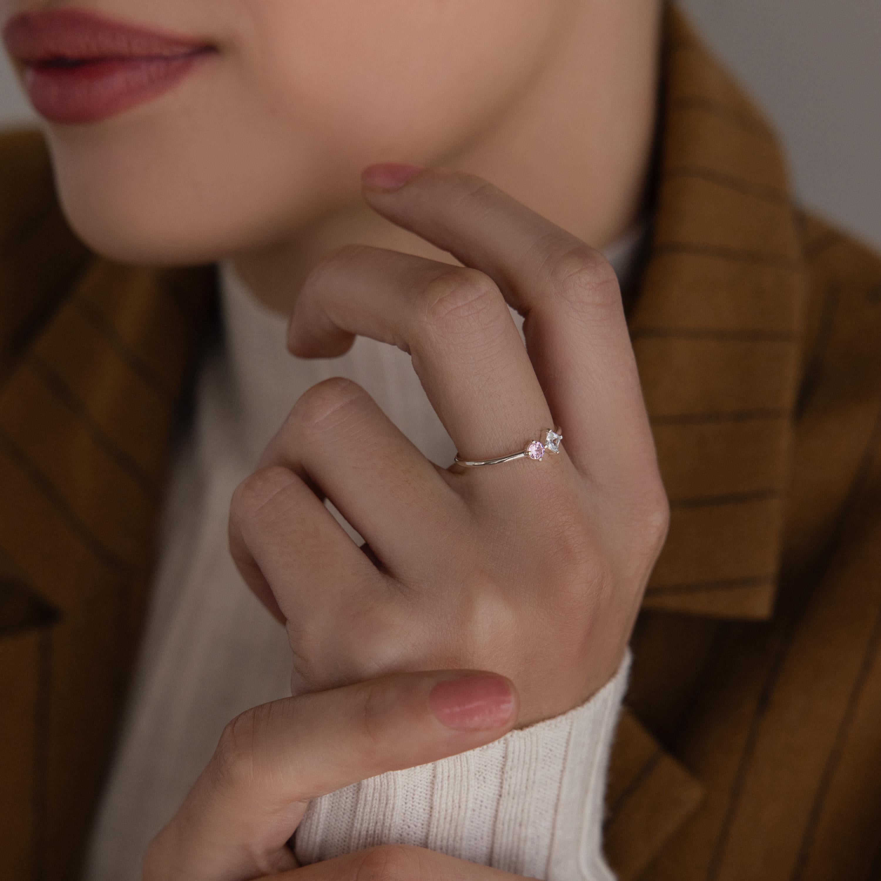 A woman wears the Toi et Moi Princess Birthstone Ring, featuring a customizable design with a small pink gemstone and a clear stone, holding her hand near her face.