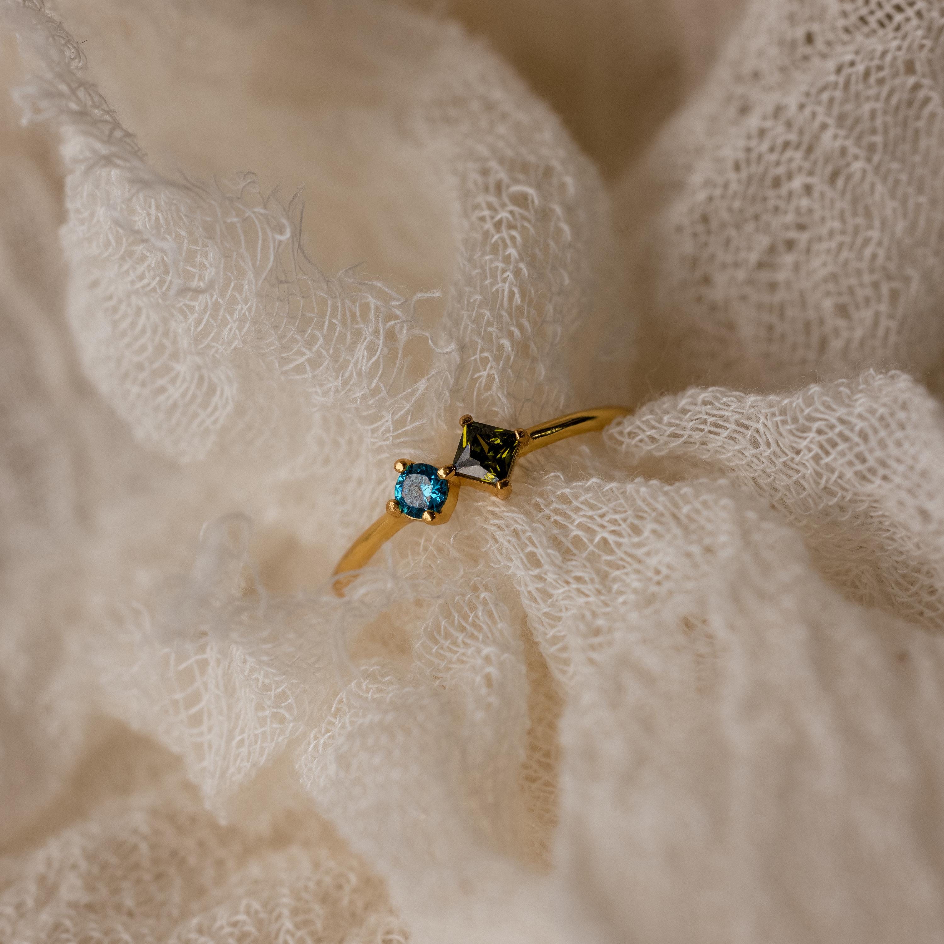 The Toi et Moi Princess Birthstone Ring, customizable with blue and green gemstones, is displayed on textured cream fabric.