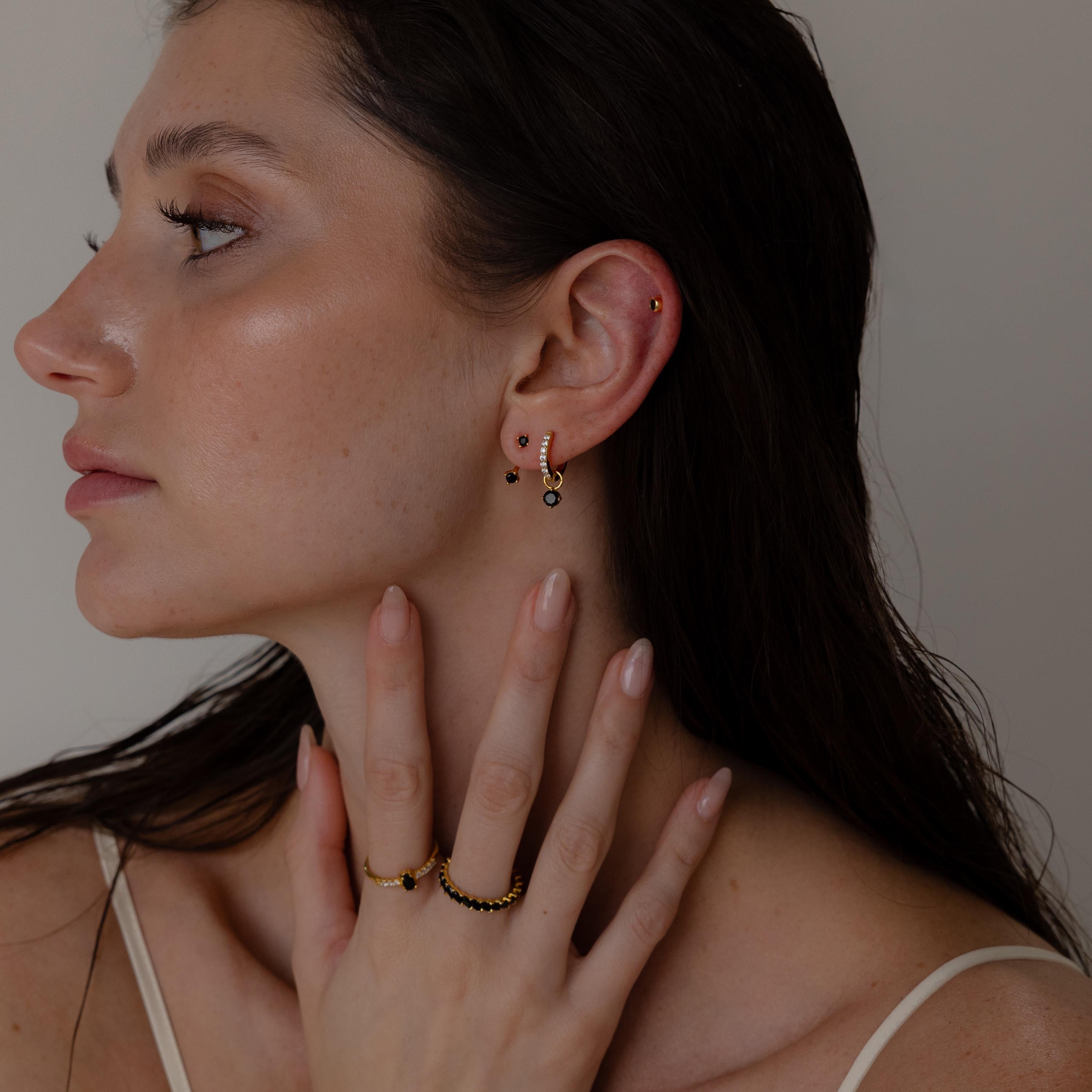 A woman with wet hair touches her neck, elegantly showcasing Black Diamond Drop Huggies earrings and rings against a neutral background.