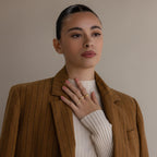 A woman in a brown pinstripe blazer and white sweater touches her chest, wearing gold rings including the Toi et Moi Infinity Birthstone Ring—an elegant symbol of love with customizable gemstones.