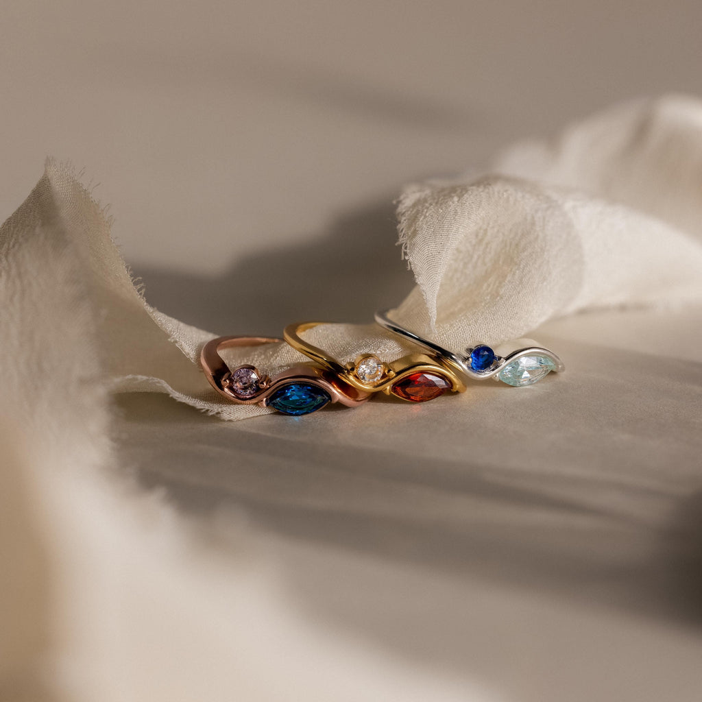 The Toi et Moi Infinity Birthstone Ring shines in gold and silver atop soft beige fabric, its customizable gemstones sparkling in gentle light—a perfect symbol of love and personal connection.