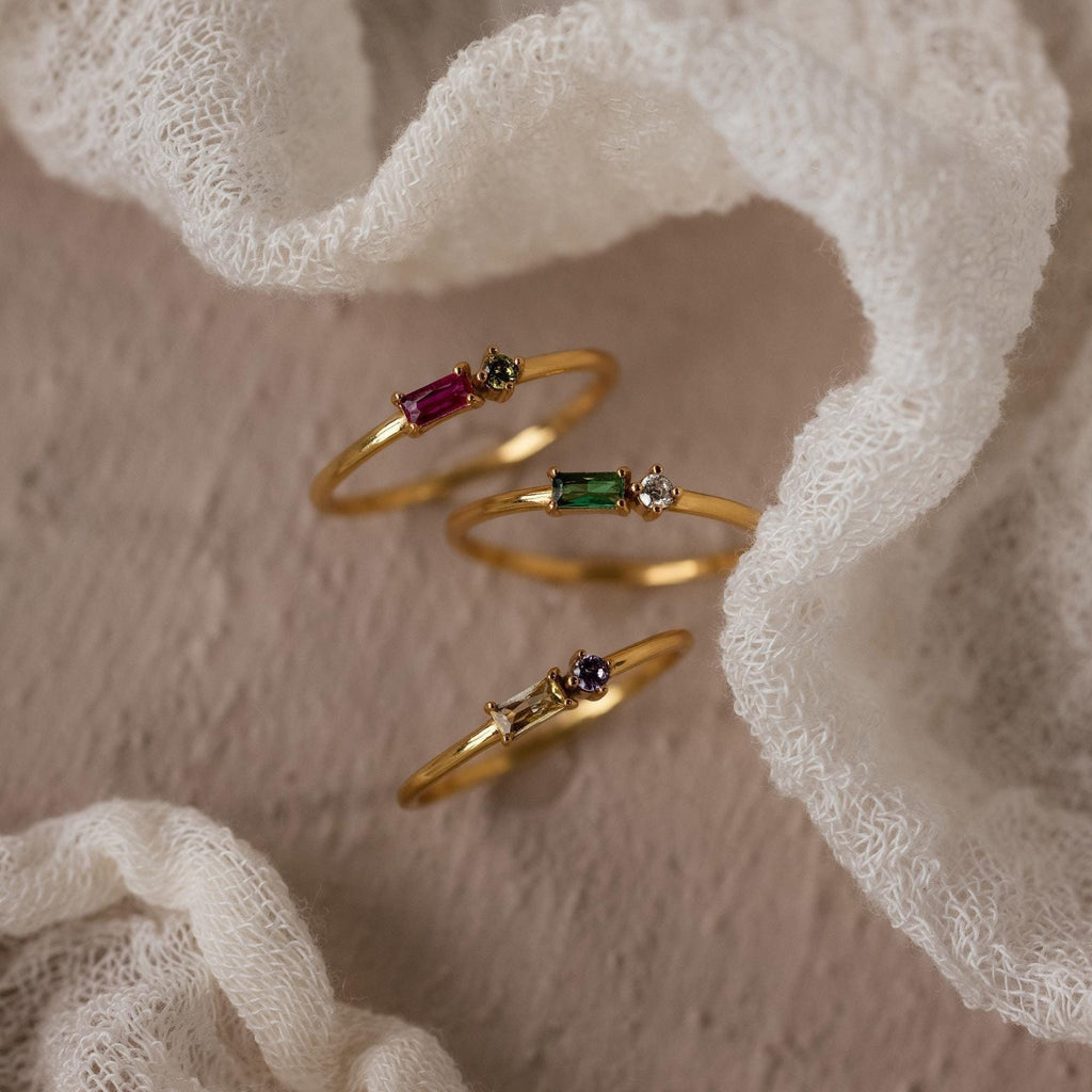 The Toi et Moi Baguette Birthstone Ring, a delicate gold piece, showcases colorful gemstones and is displayed on a soft, textured white fabric background.