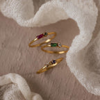 The Toi et Moi Baguette Birthstone Ring, a delicate gold piece, showcases colorful gemstones and is displayed on a soft, textured white fabric background.