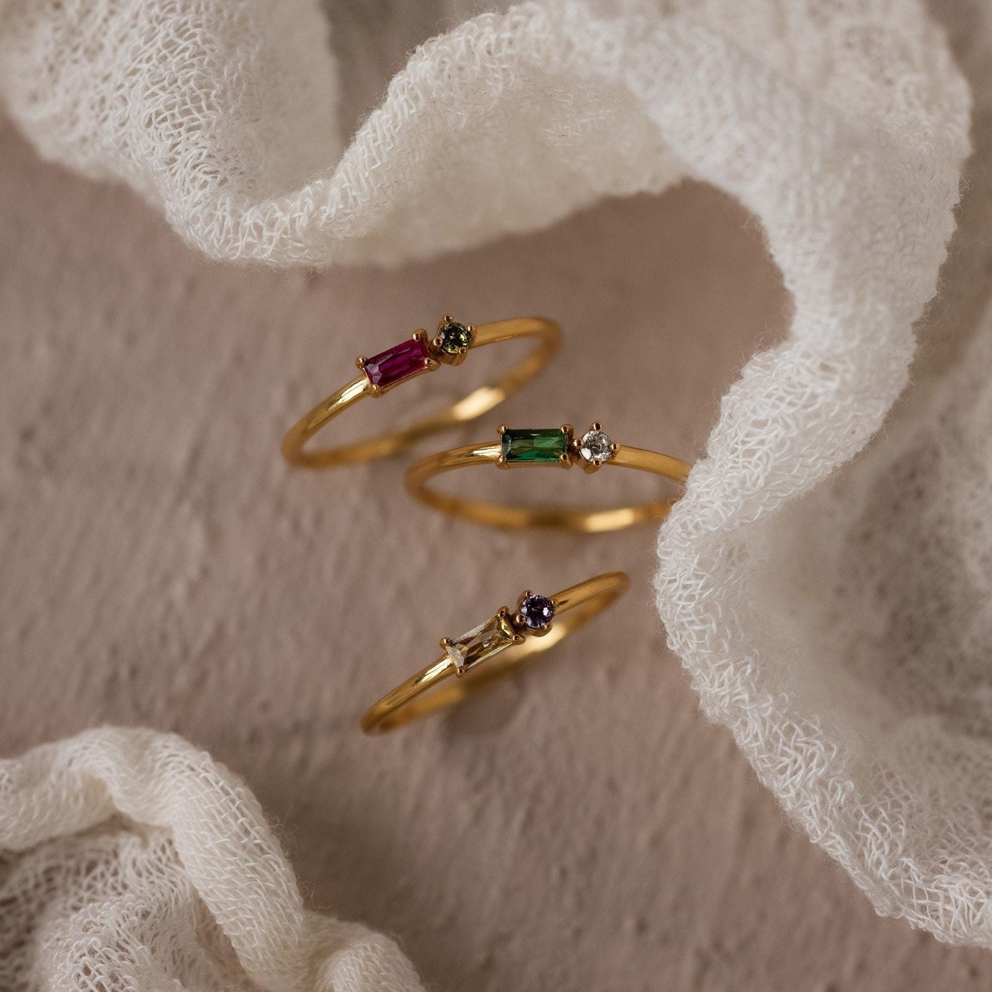 The Toi et Moi Baguette Birthstone Ring, a delicate gold piece, showcases colorful gemstones and is displayed on a soft, textured white fabric background.