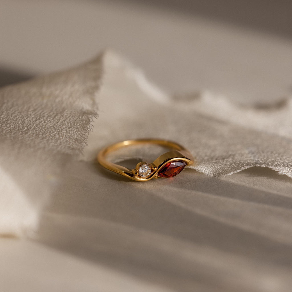 The Toi et Moi Infinity Birthstone Ring features a red marquise gemstone and a round clear stone, displayed on textured cream fabric—a perfect token of love.