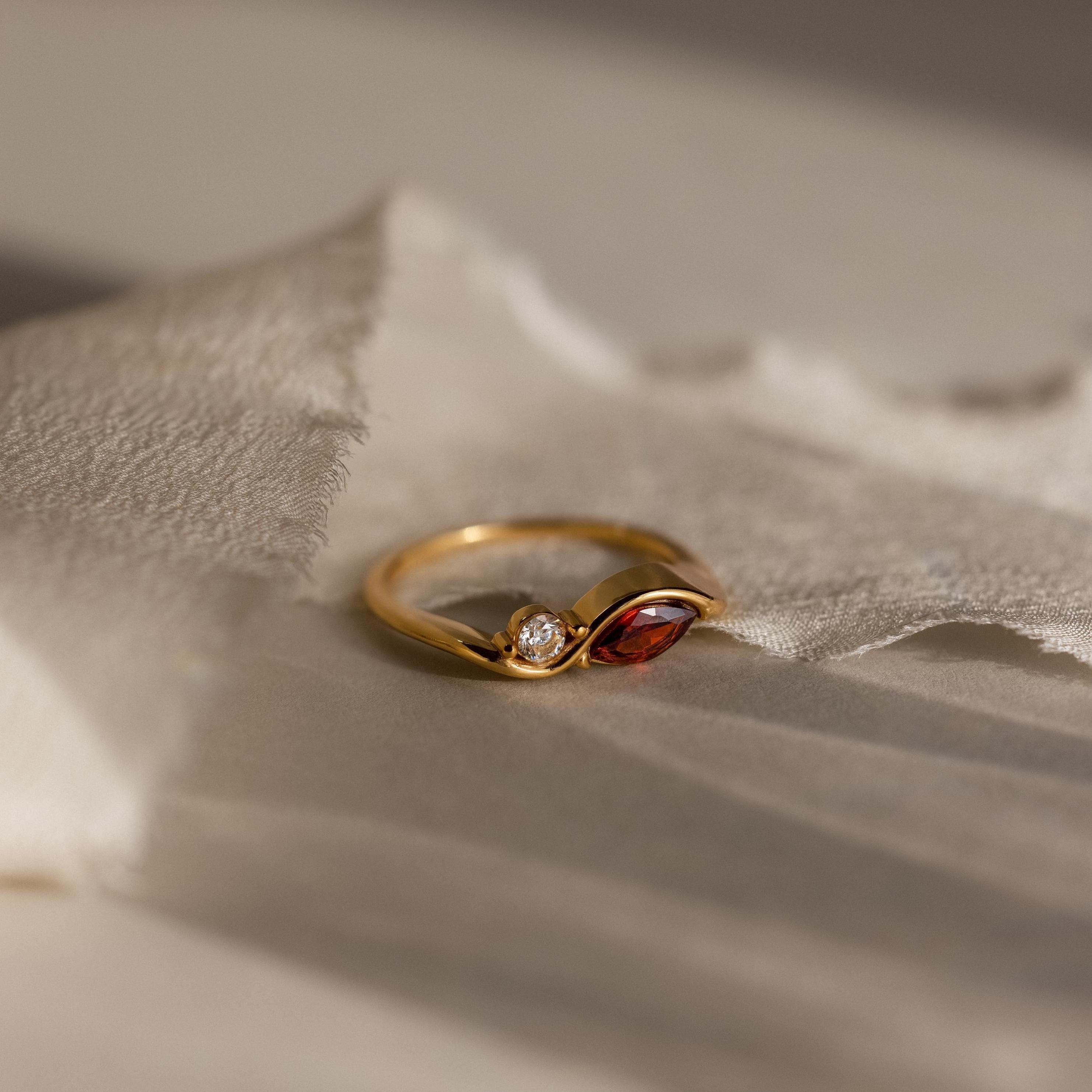 The Toi et Moi Infinity Birthstone Ring features a red marquise gemstone and a round clear stone, displayed on textured cream fabric—a perfect token of love.