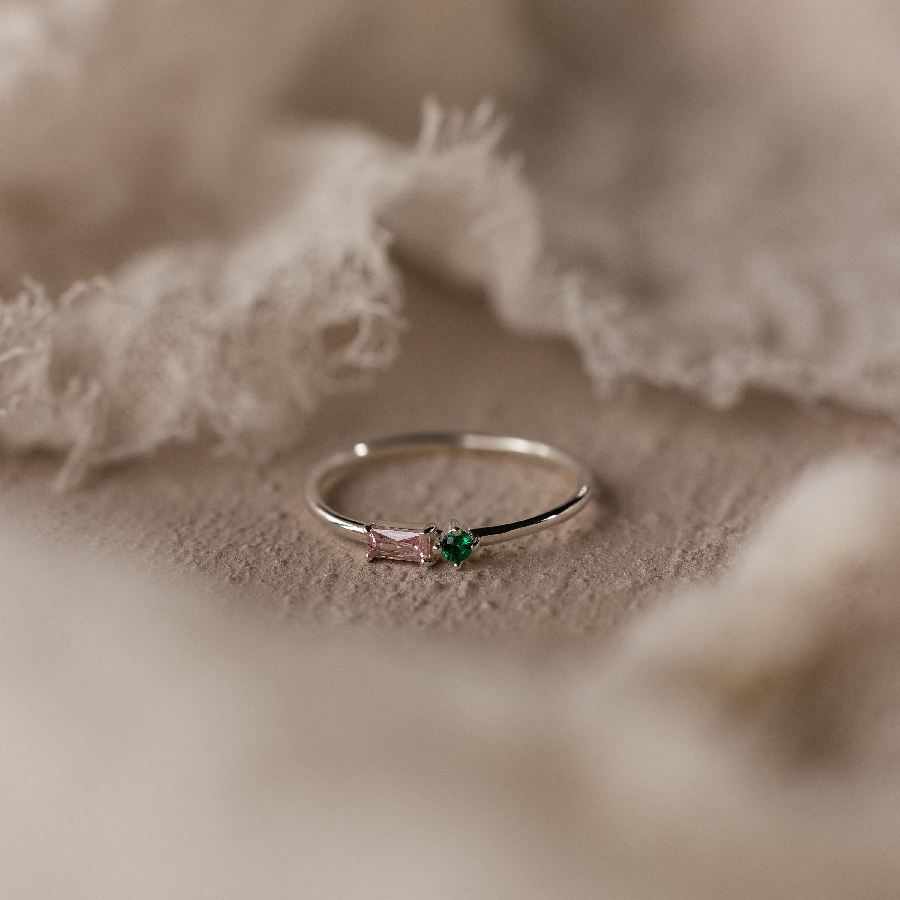 The Toi et Moi Baguette Birthstone Ring, featuring pink and green gemstones, is displayed on a soft beige fabric background.