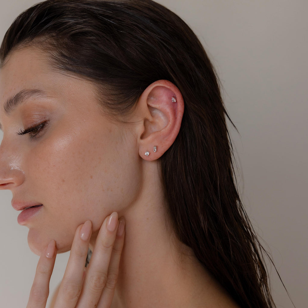 A woman with wet hair showcases multiple ear piercings adorned with the Mixed Diamond Flat Back Stud Set, gently touching her chin against a neutral background.