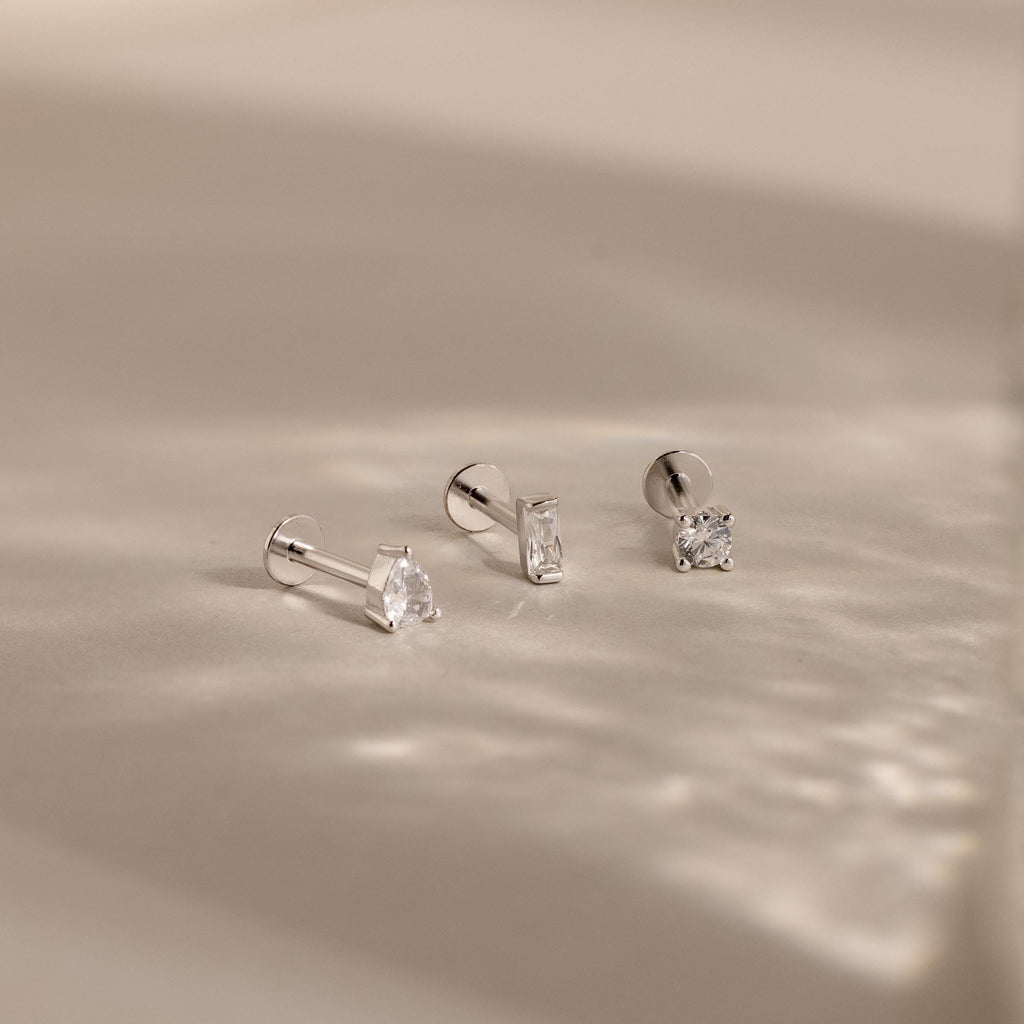 The Mixed Diamond Flat Back Stud Set features three minimalist silver stud earrings with clear gemstones, displayed on a soft, light beige surface.