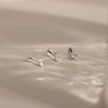 The Mixed Diamond Flat Back Stud Set features three minimalist silver stud earrings with clear gemstones, displayed on a soft, light beige surface.