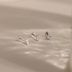 The Mixed Diamond Flat Back Stud Set features three minimalist silver stud earrings with clear gemstones, displayed on a soft, light beige surface.