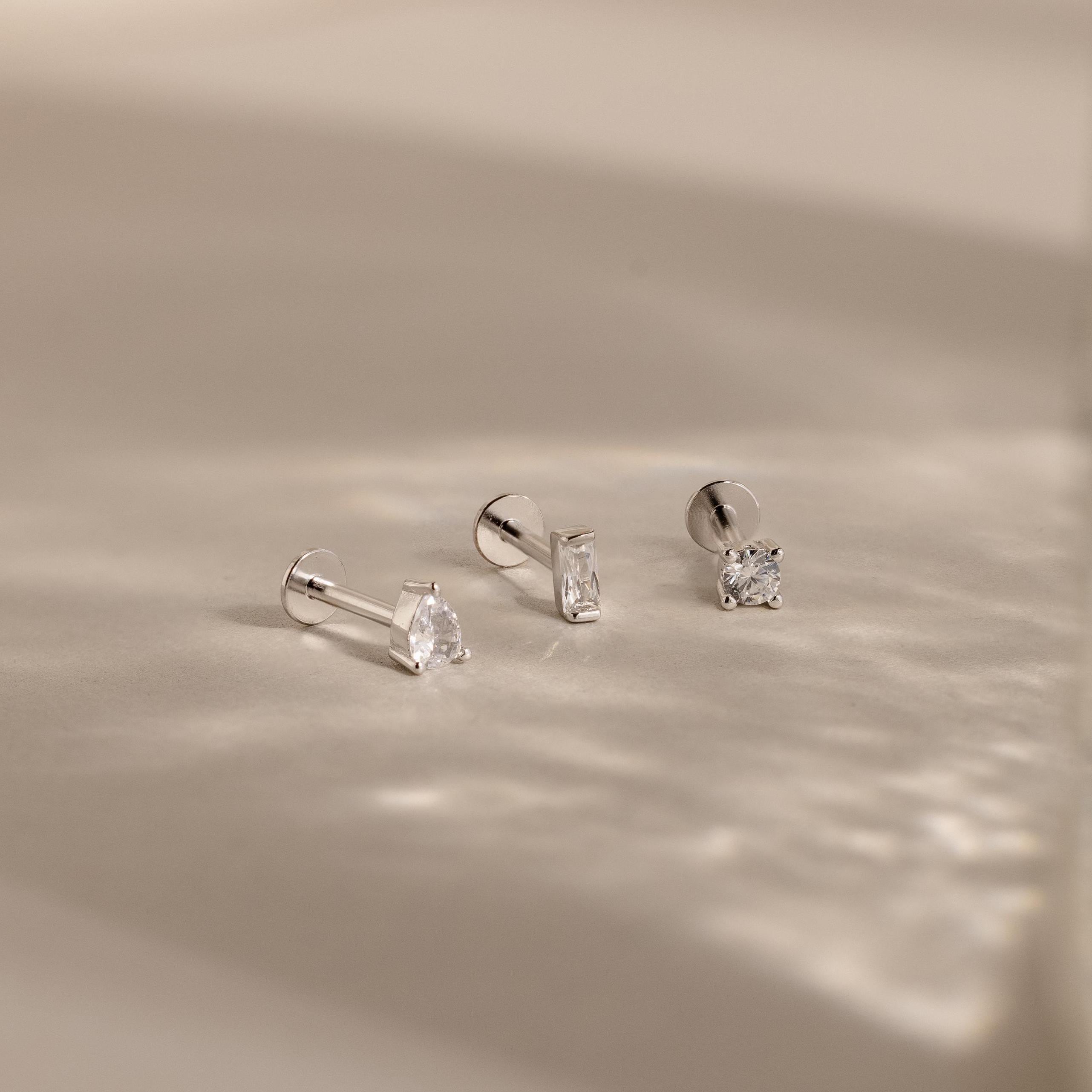 The Mixed Diamond Flat Back Stud Set features three minimalist silver stud earrings with clear gemstones, displayed on a soft, light beige surface.