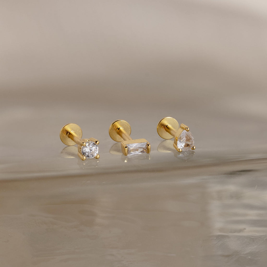The Mixed Diamond Flat Back Stud Set includes three gold earrings—round, rectangular, and teardrop-shaped—with clear CZ stones, ideal for an elegant look and displayed on a reflective, light-colored surface.