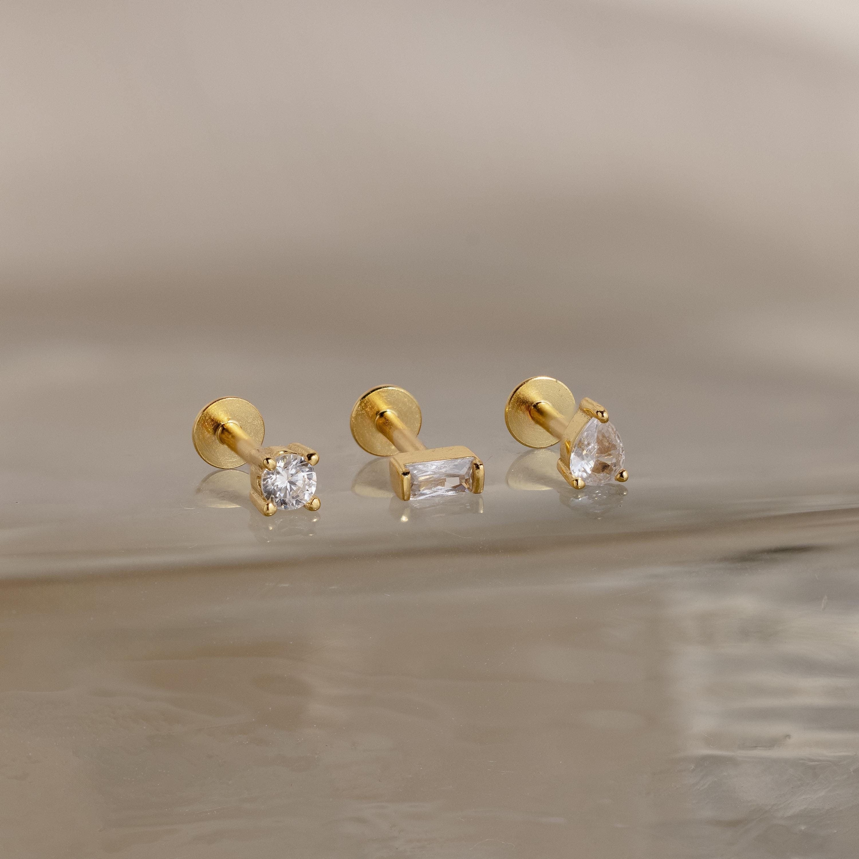 The Mixed Diamond Flat Back Stud Set includes three gold earrings—round, rectangular, and teardrop-shaped—with clear CZ stones, ideal for an elegant look and displayed on a reflective, light-colored surface.