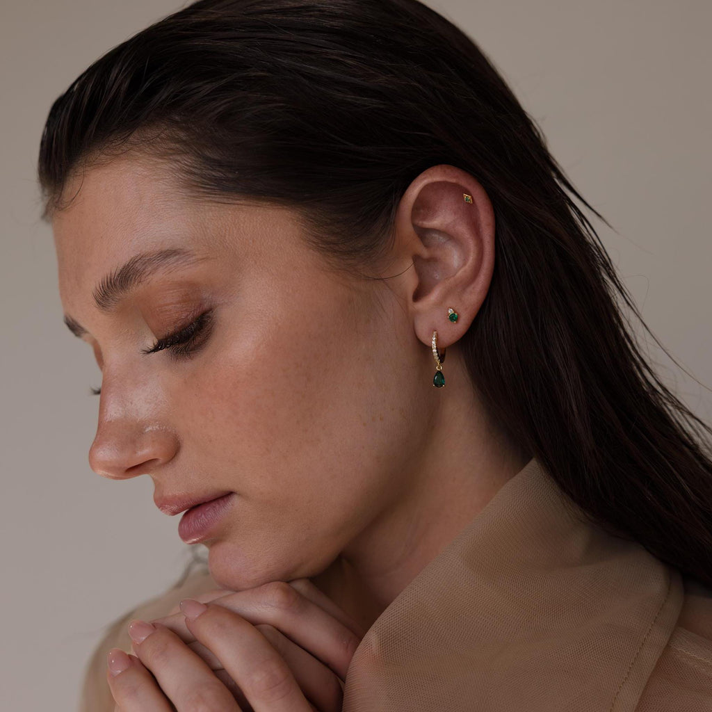 A woman wearing multiple earrings including a gold geometric flat back stud on her upper ear, styled with green gemstone drop and stud earrings.