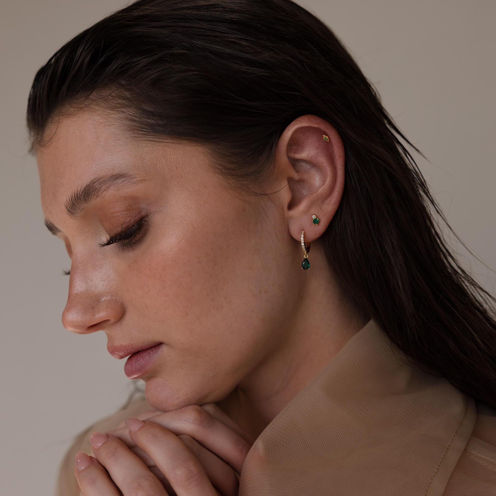 A woman wearing multiple earrings including a gold geometric flat back stud on her upper ear, styled with green gemstone drop and stud earrings.