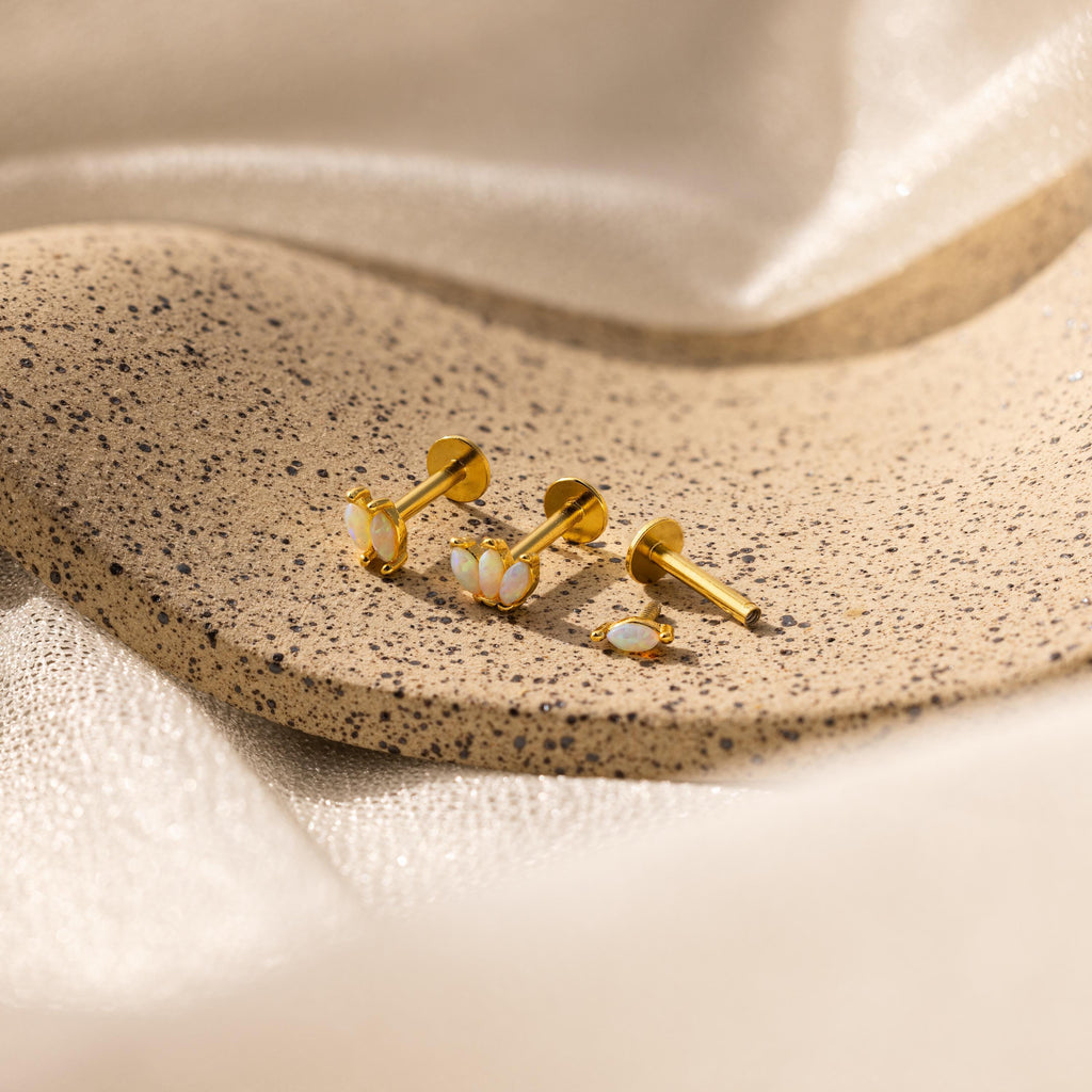 Gold earrings with white stones, like our Marquise Opal Flat Back Studs, displayed on a speckled stone surface with soft beige fabric in the background—a perfect gift for her.