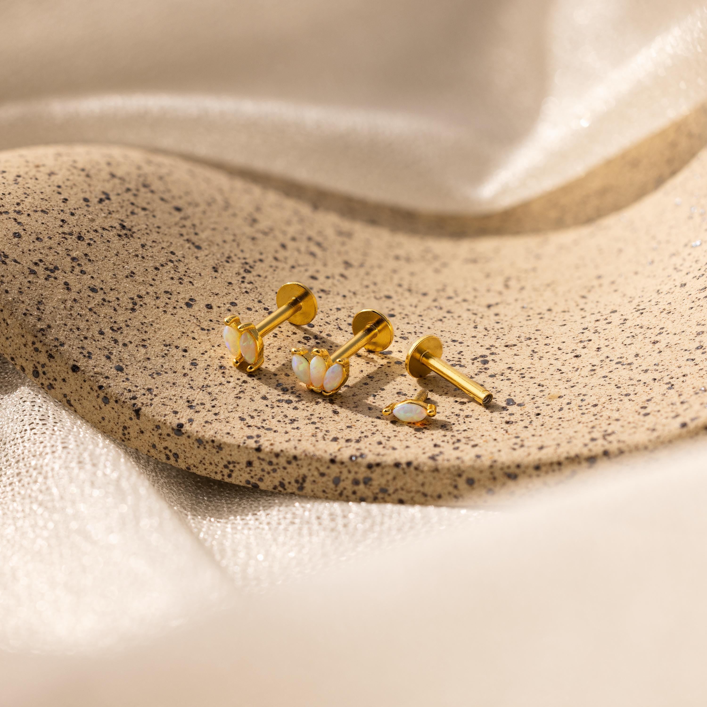 Gold earrings with white stones, like our Marquise Opal Flat Back Studs, displayed on a speckled stone surface with soft beige fabric in the background—a perfect gift for her.