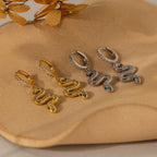 Pave Snake Charm Hoops with gemstone eyes in gold and silver are displayed on a beige, curved surface.