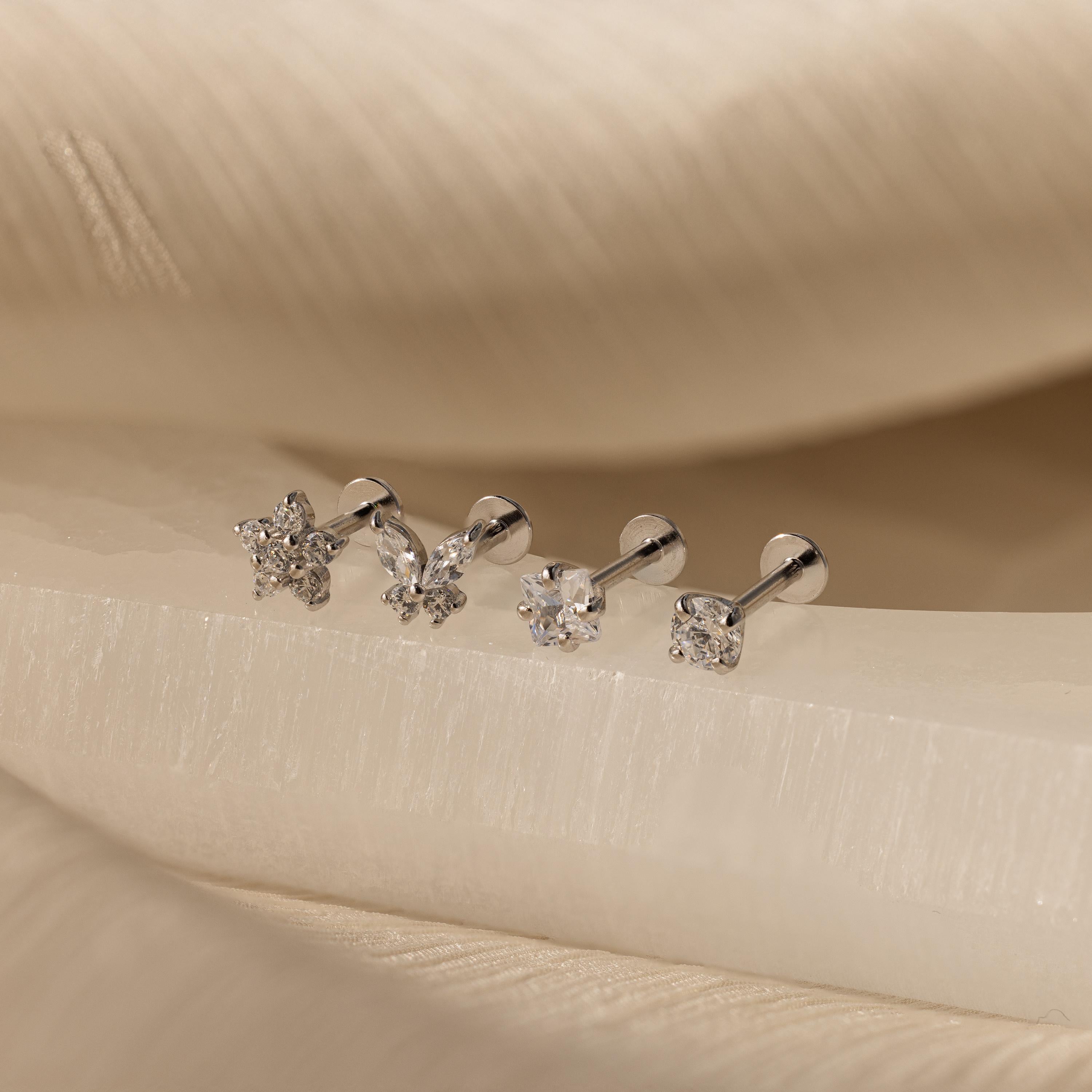 The Pave Assortment Flat Back Set features four earrings, each with a distinct crystal shape, displayed on cream fabric. This elegant set brings sparkle and style to any look.
