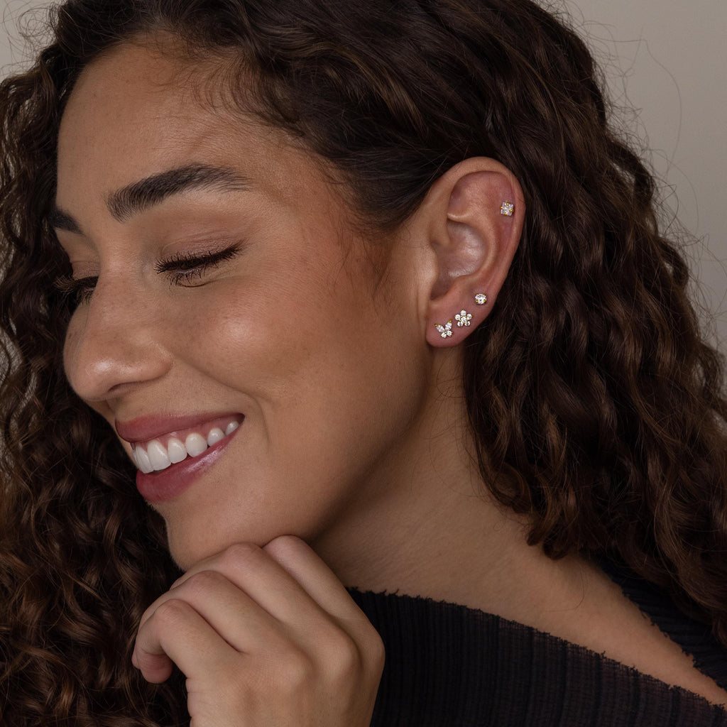 A smiling woman with curly hair models the Pave Assortment Flat Back Set, featuring multiple silver and pave diamond flat-back studs on her ear, paired with a dark top.