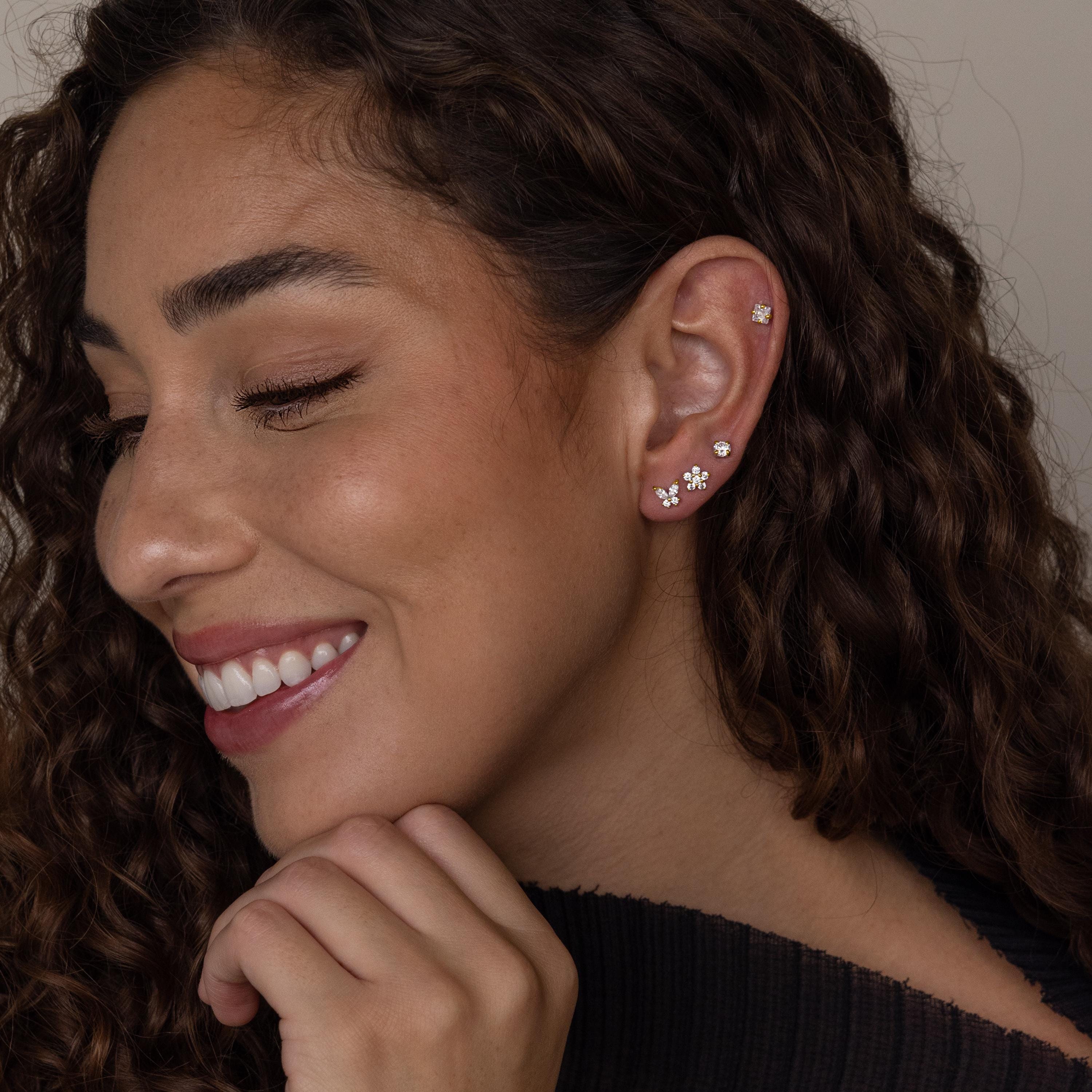 A smiling woman with curly hair models the Pave Assortment Flat Back Set, featuring multiple silver and pave diamond flat-back studs on her ear, paired with a dark top.
