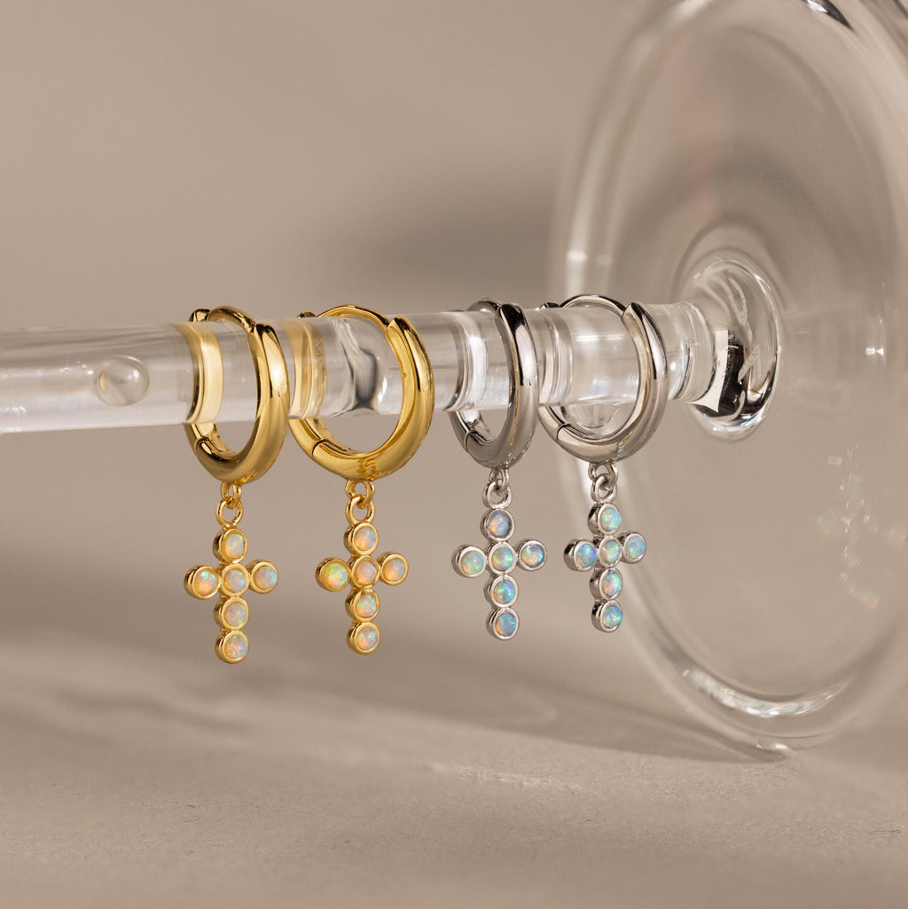 Showcase your style with Opal Cross Huggies—two pairs of huggie earrings with opal cross pendants, one in gold and one in silver, elegantly displayed on a glass stem.