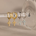 Showcase your style with Opal Cross Huggies—two pairs of huggie earrings with opal cross pendants, one in gold and one in silver, elegantly displayed on a glass stem.