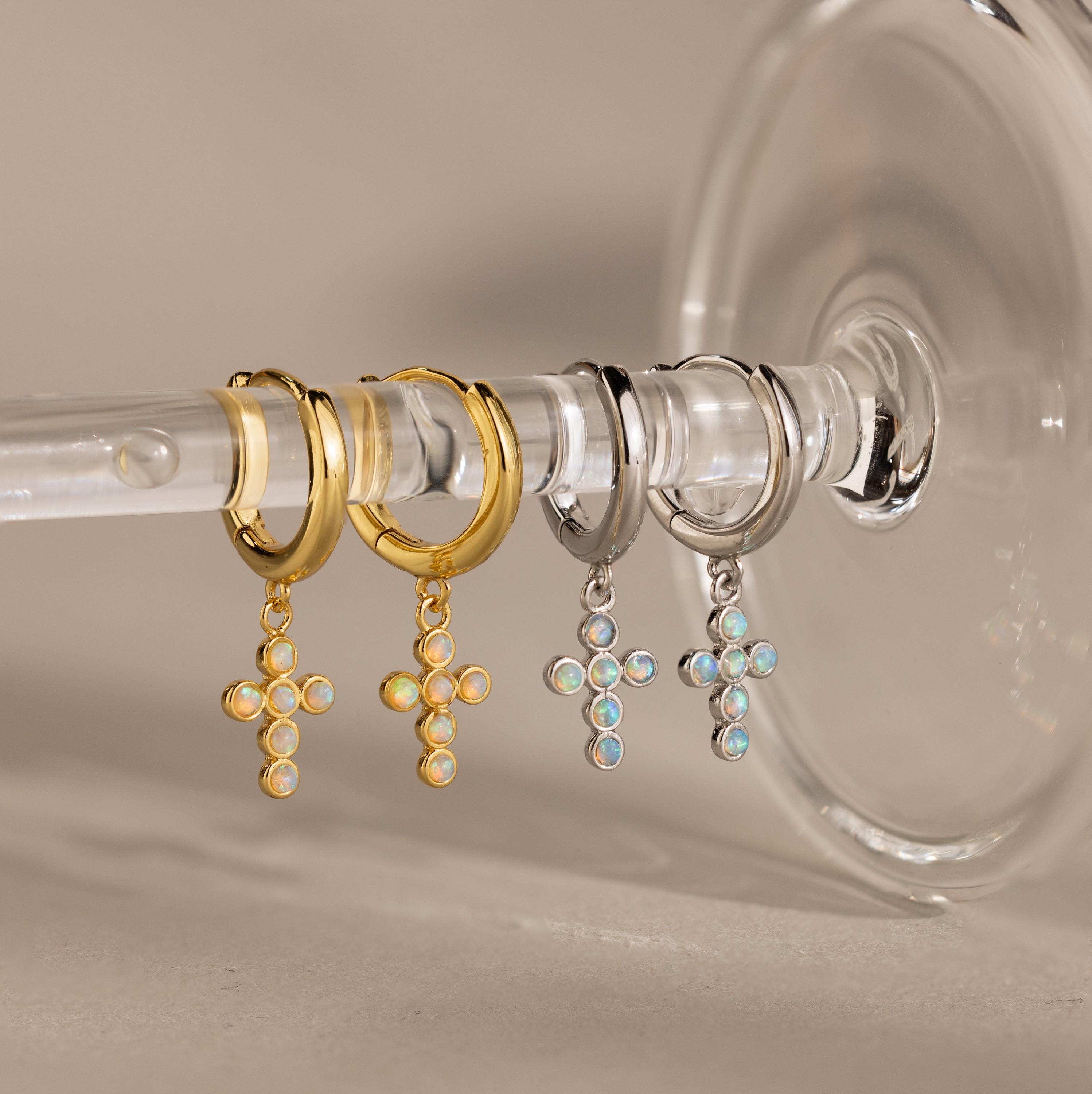 Showcase your style with Opal Cross Huggies—two pairs of huggie earrings with opal cross pendants, one in gold and one in silver, elegantly displayed on a glass stem.