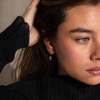 A woman in a black top touches her hair and gazes to the side, wearing delicate Opal Cross Huggies earrings.