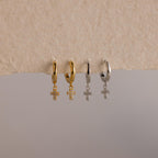 Two gold and two silver Pave Cross Huggies, each with a delicate dangling cross pendant, are beautifully displayed on a textured surface.