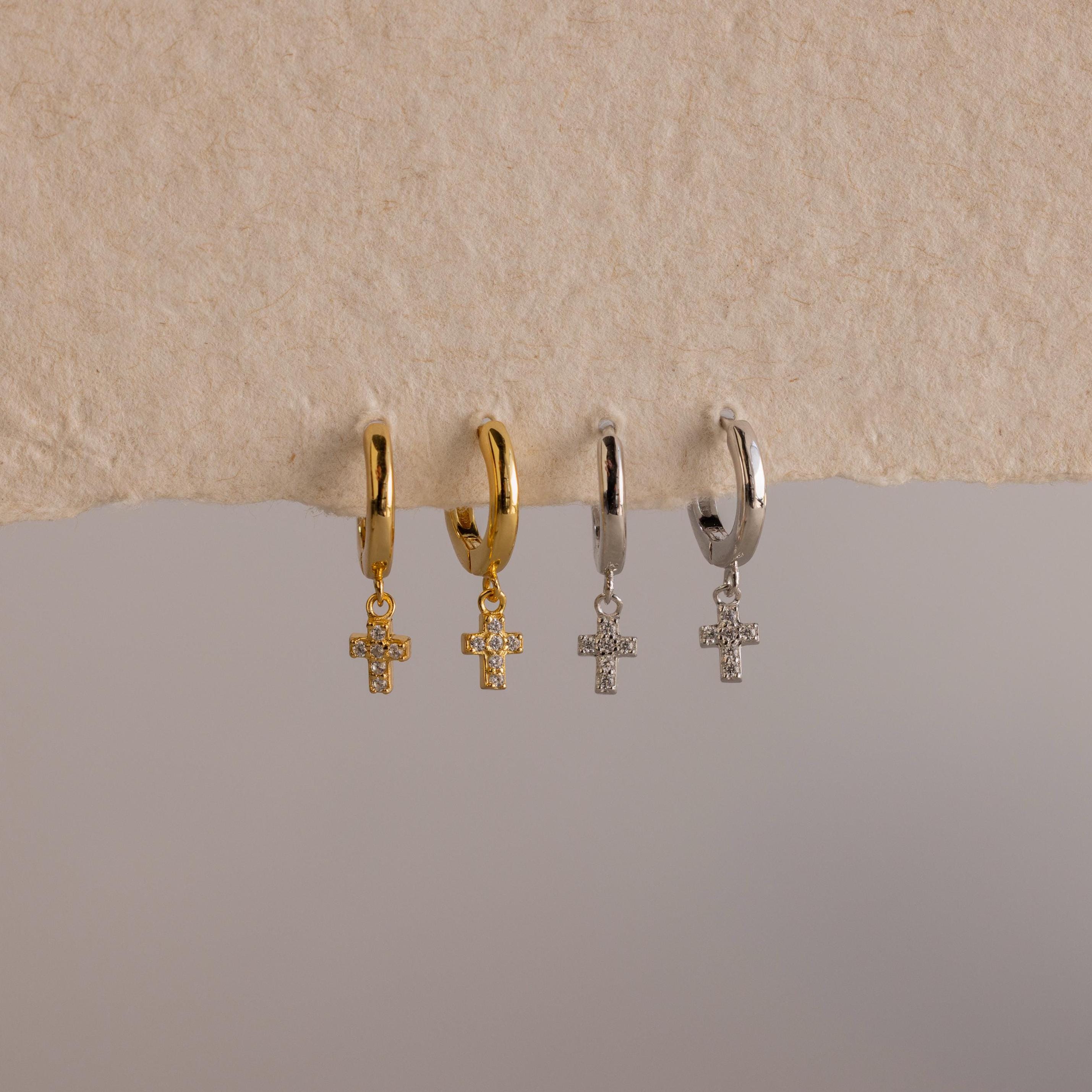 Two gold and two silver Pave Cross Huggies, each with a delicate dangling cross pendant, are beautifully displayed on a textured surface.