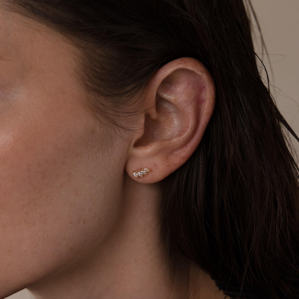 A close-up shows a woman's ear adorned with the Marquise Diamond Climbers—small, delicate gold earrings featuring multiple leaf-shaped stones, ideal for elegant wedding jewelry.