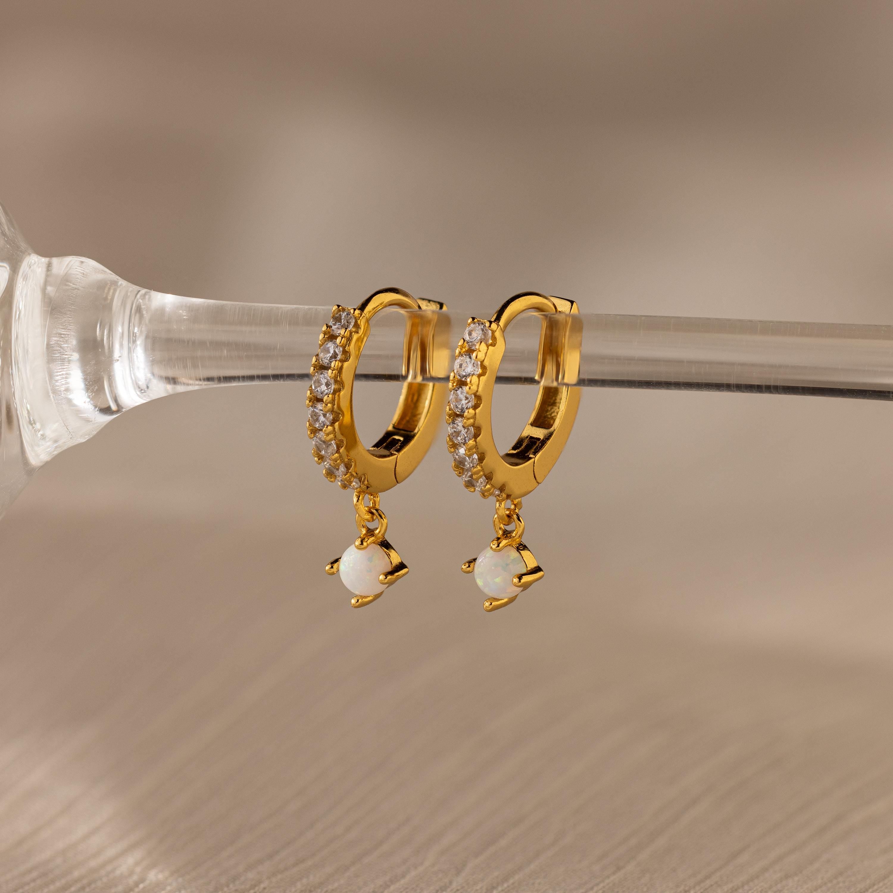 Pave Opal Drop Huggies—gold hoop earrings with sparkling white stones and dangling pearls, reminiscent of opal earrings, displayed on a clear glass rod.