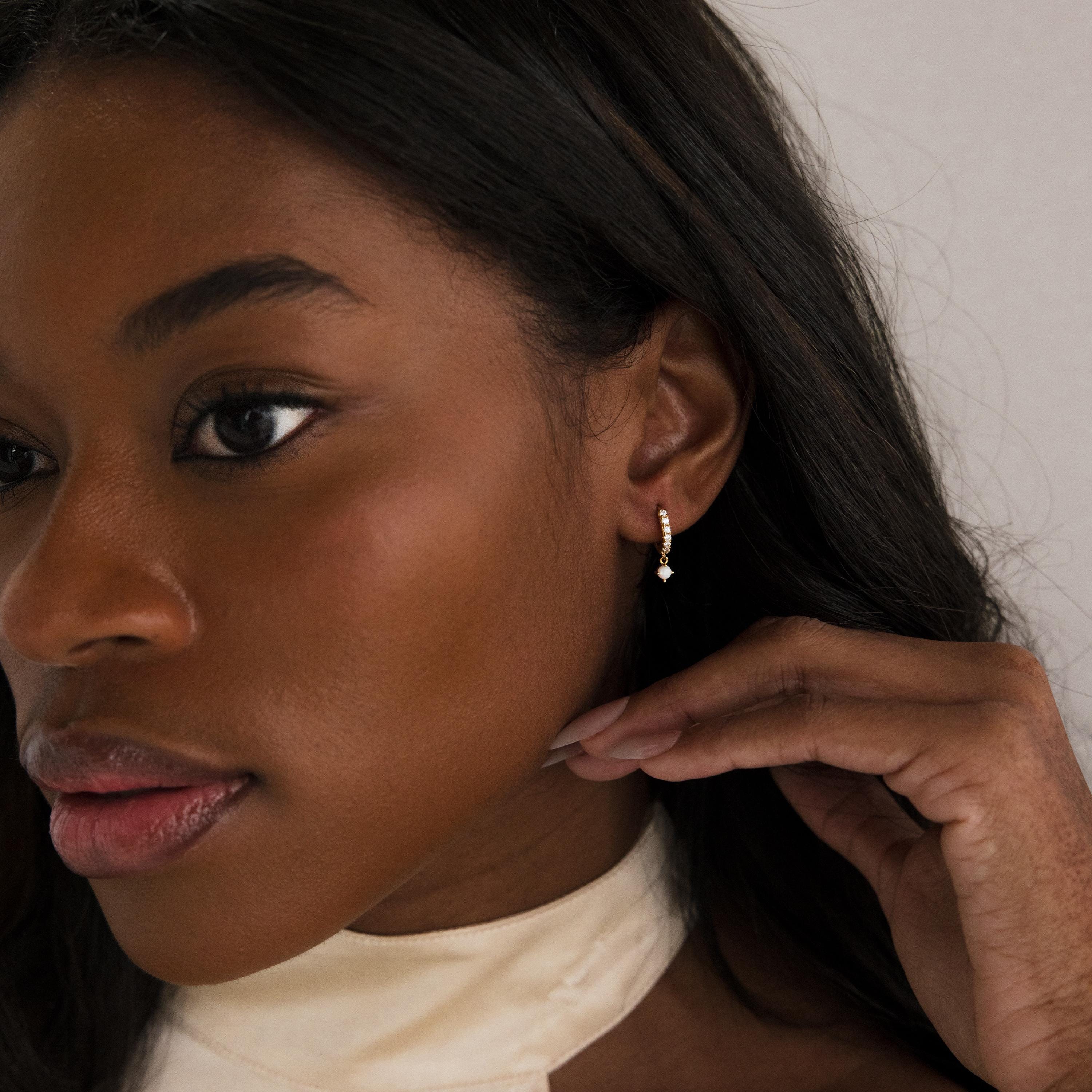 Woman with long dark hair touches her ear while wearing Pave Opal Drop Huggies against a neutral background.
