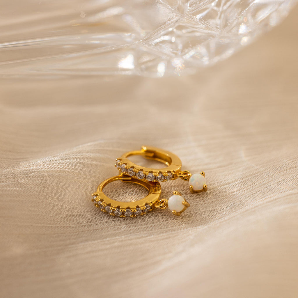 Pave Opal Drop Huggies rest on soft beige fabric under gentle light, showcasing gold hoops with pavé-set opals and elegant pearl drops.