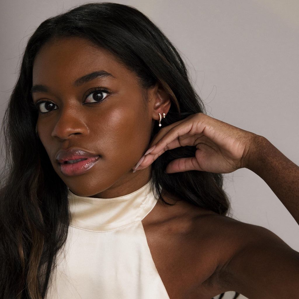 A woman with long dark hair in a cream halter top touches her ear, highlighting the Pave Opal Drop Huggies against a neutral background.
