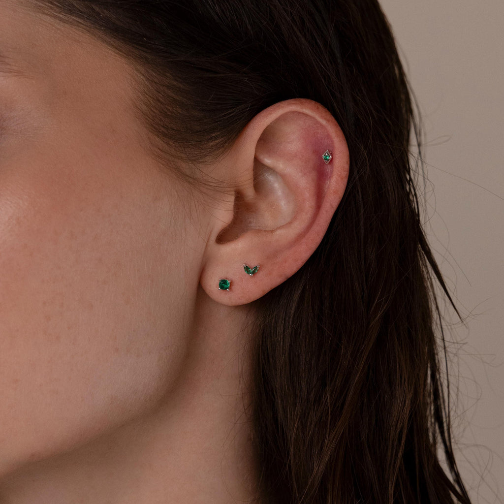 Close-up of a woman's ear wearing the Emerald Flat Back Earring Set, featuring three small emerald studs in the lobe and one screw-back earring in the cartilage.