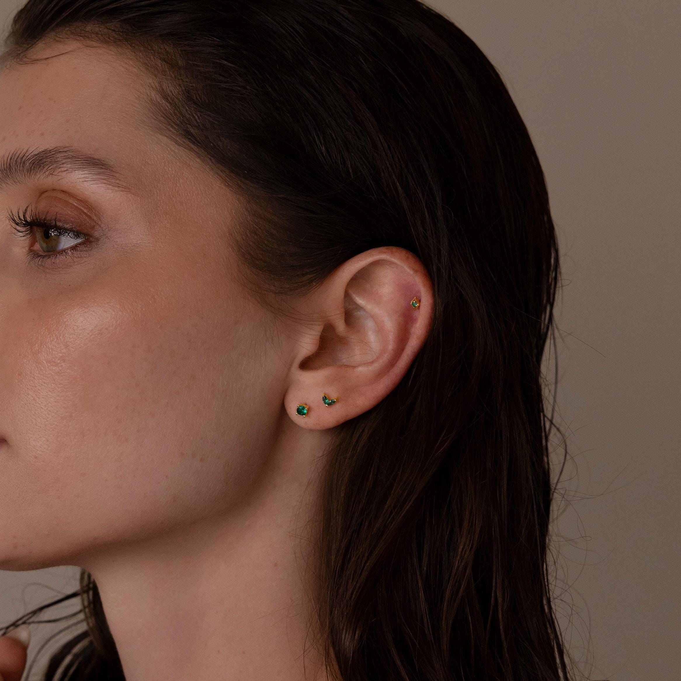 Close-up of a woman's ear adorned with the Emerald Flat Back Earring Set, featuring three small green emerald earrings—birthstone studs styled on the lobe and cartilage.