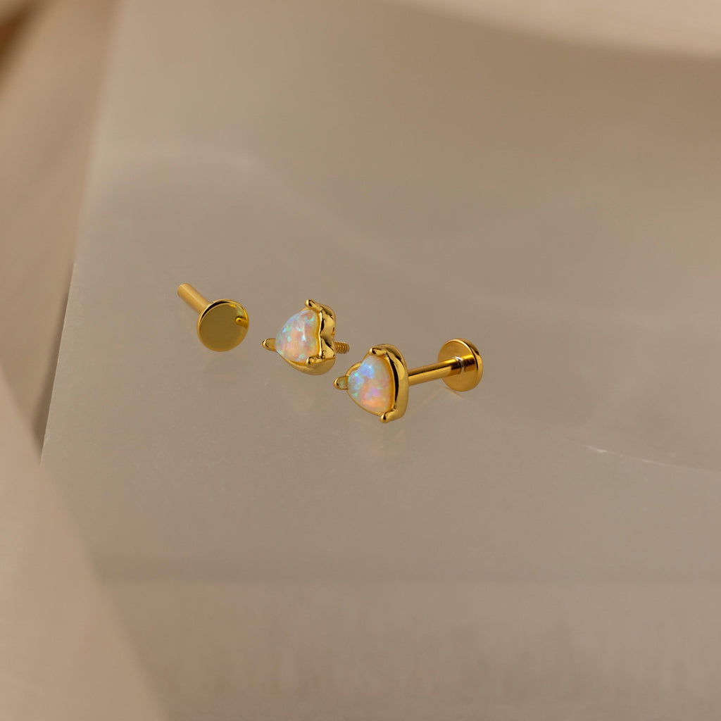 The Opal Heart Flat Back Studs, featuring iridescent heart-shaped stones, are displayed on a soft beige surface.
