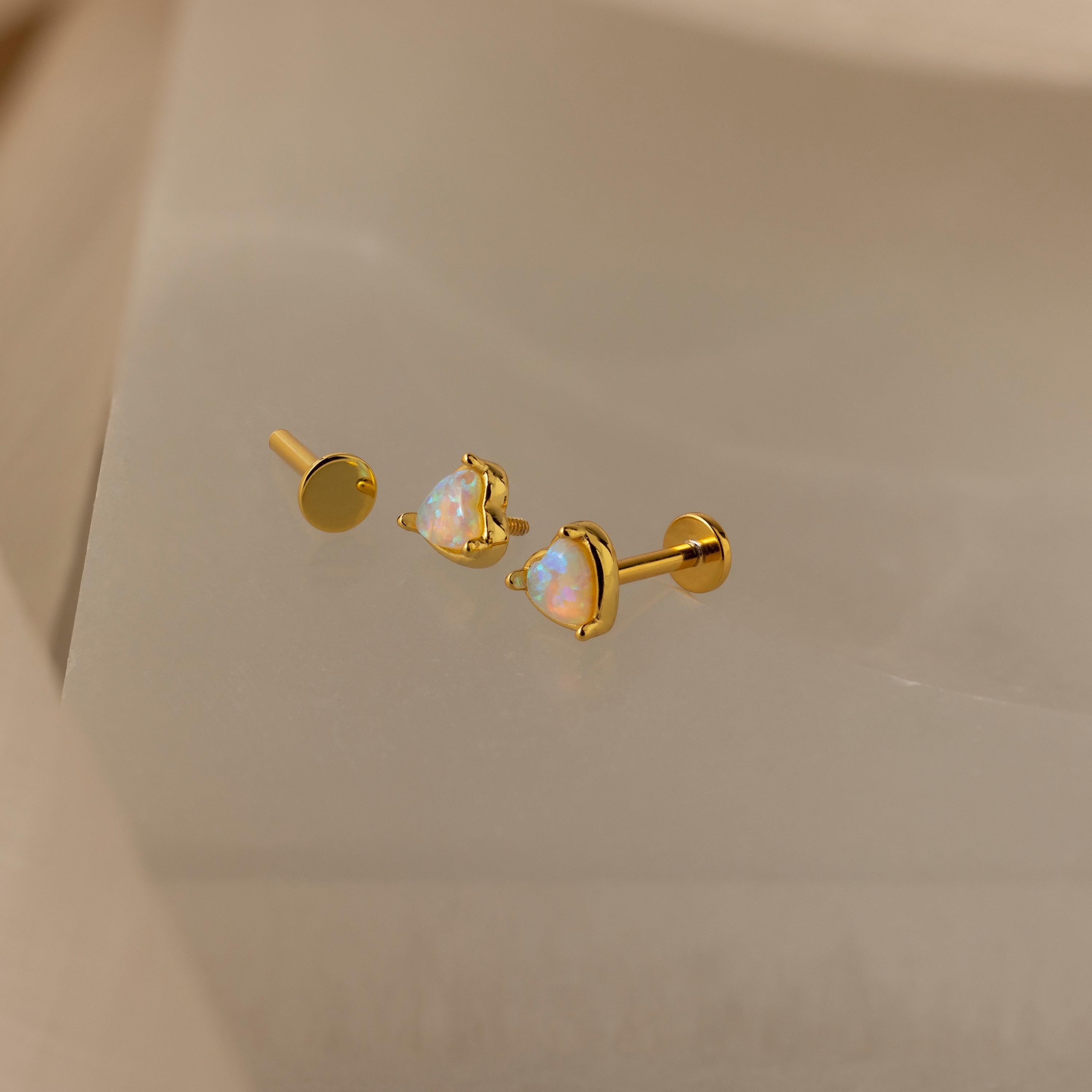 The Opal Heart Flat Back Studs, featuring iridescent heart-shaped stones, are displayed on a soft beige surface.